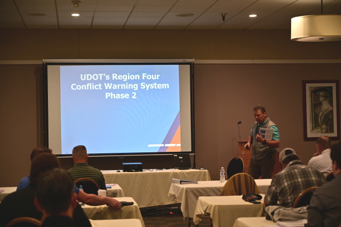 A man presents beside a projector screen showing a title slide with text UDOT's Region Four Conflict Warning System Phase 2.